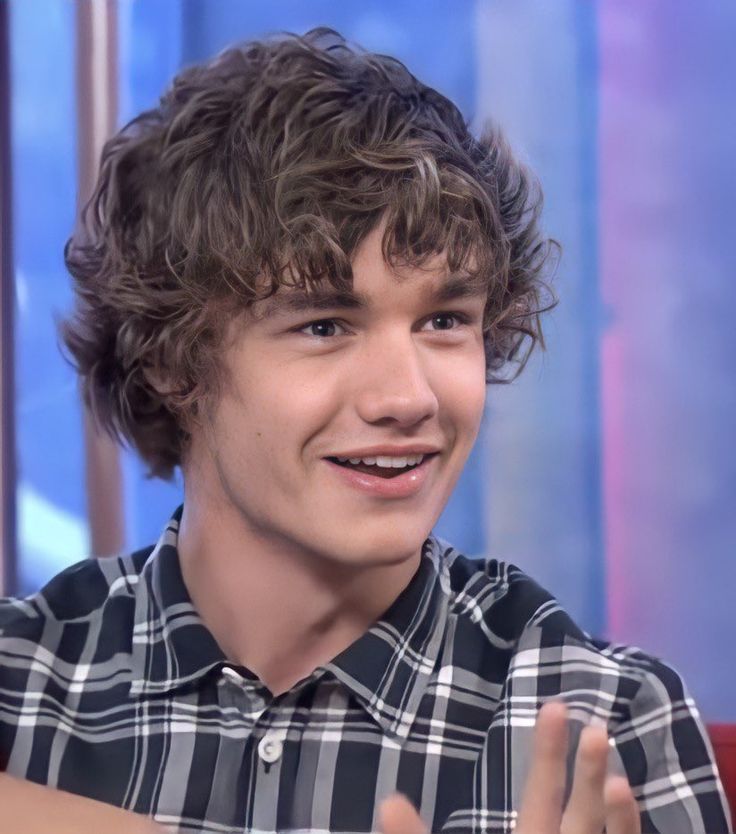 a young man with curly hair sitting in front of a red chair and smiling at the camera