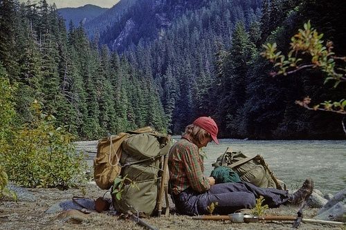 two people sitting on the ground next to a river with mountains in the background and trees