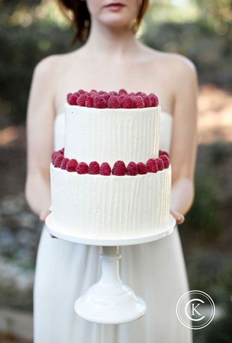 a woman holding a white cake with raspberries on the top and bottom tier