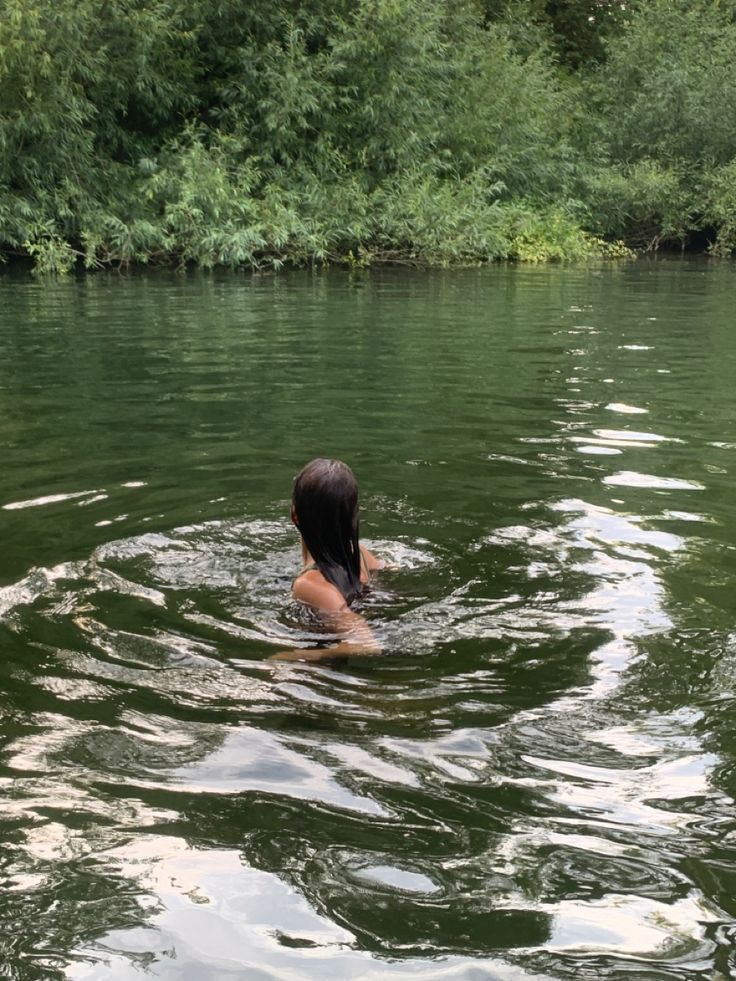 a woman is swimming in the water with trees in the backgrouds and bushes behind her