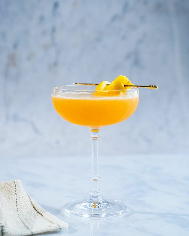 an orange cocktail in a coupe glass on a marble table with a cloth and napkin