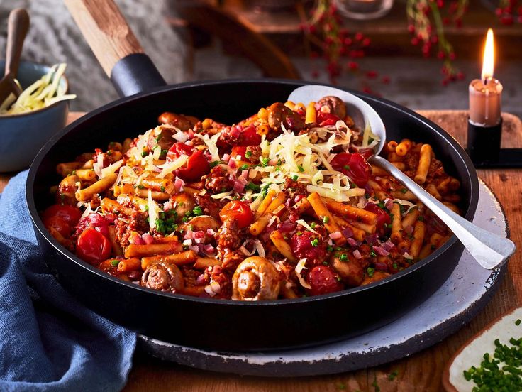 a skillet filled with pasta and vegetables on top of a wooden table next to a lit candle