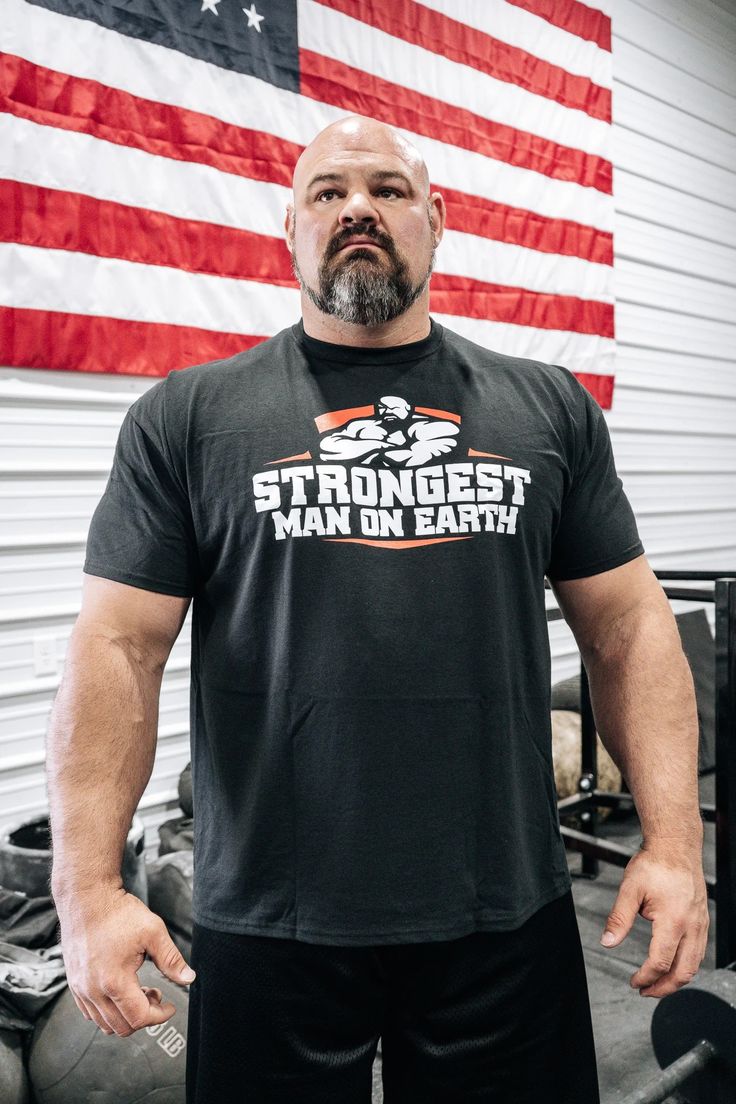 a man standing in front of an american flag wearing a black shirt that says, strongest man on earth