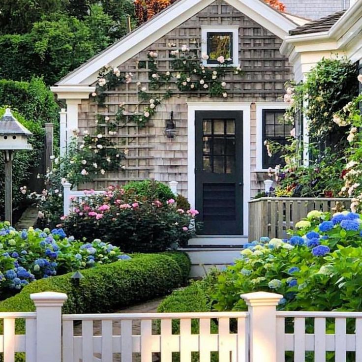 a house with lots of flowers and bushes around it's front door, next to a white picket fence