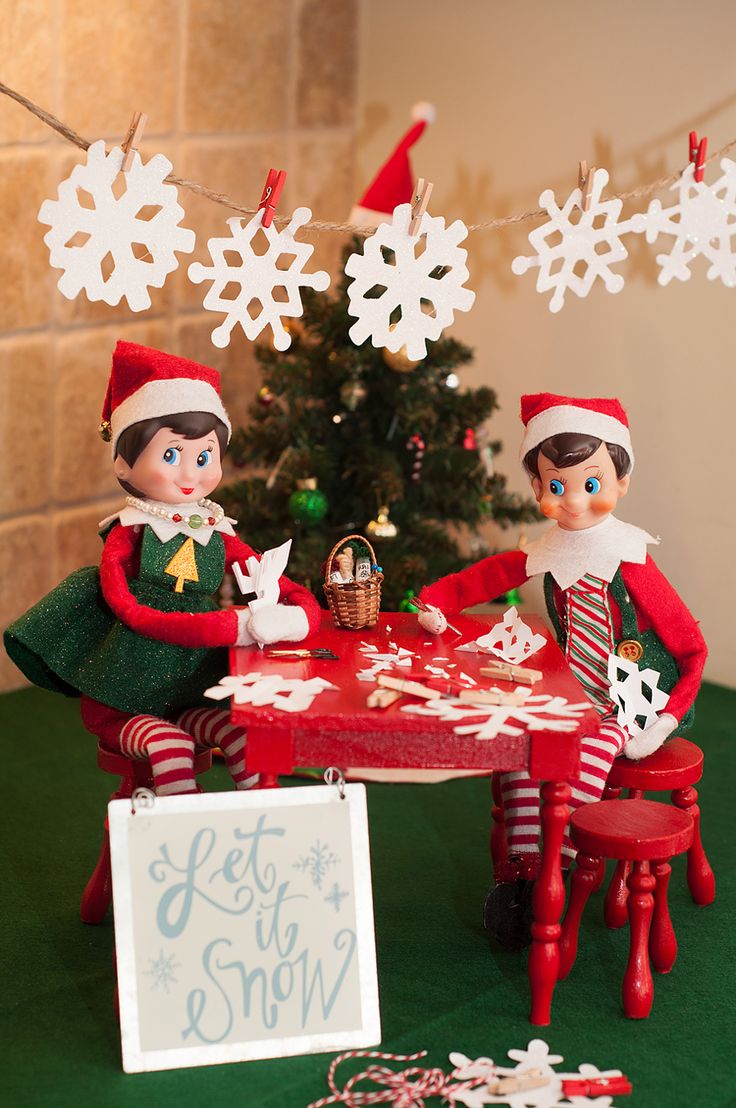 two elfs are sitting at a table near a christmas tree with snowflakes