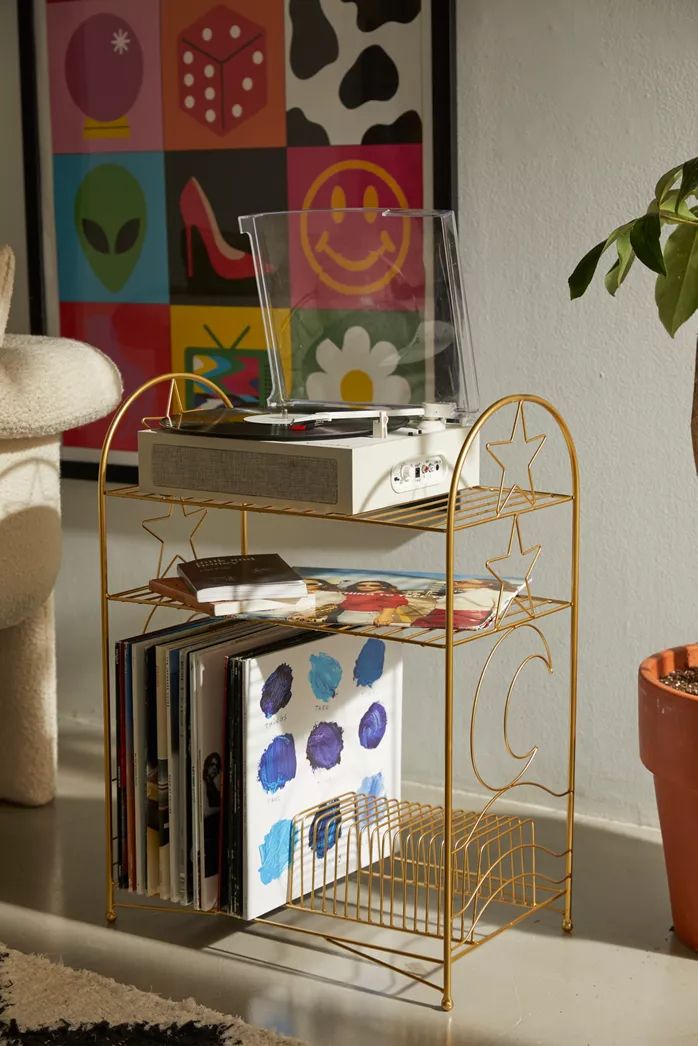 a gold metal shelf with cd's and magazines on it next to a potted plant