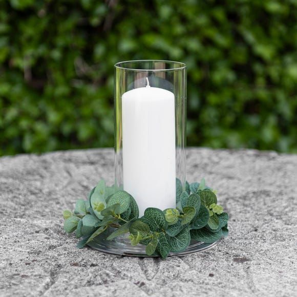 a white candle sitting on top of a glass vase filled with leaves and greenery