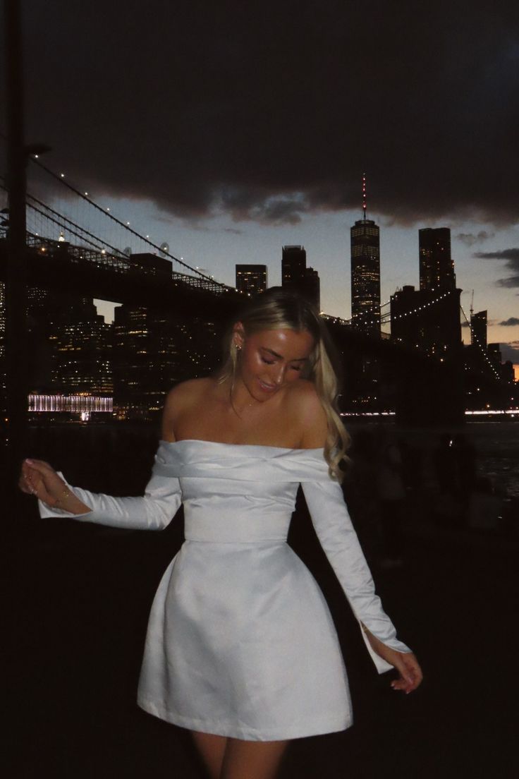 a woman in a white dress is walking by the water at night with city lights behind her