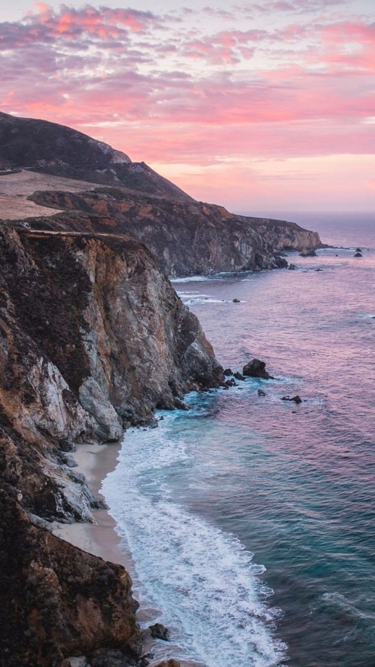 an ocean cliff with waves crashing on the shore and pink clouds in the sky above