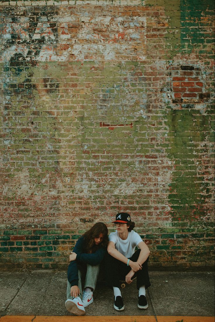 two people sitting on the ground next to a brick wall