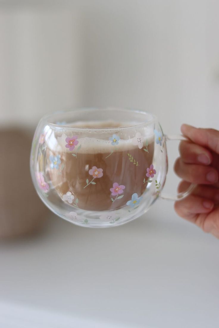 a hand holding a glass with liquid in it and flowers painted on the side,