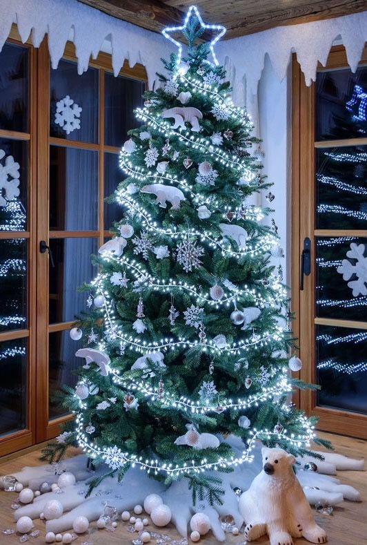 a christmas tree with white lights and decorations in front of two windows, one dog laying on the floor