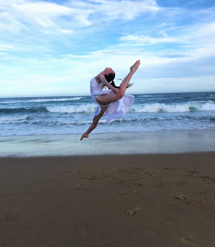 a woman in white dress jumping into the air on beach next to ocean with waves