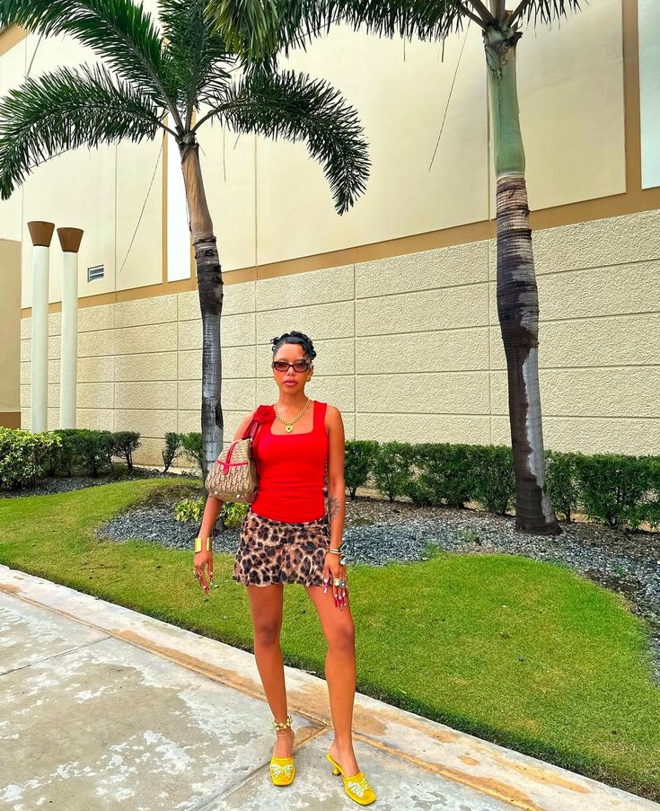 a woman in red shirt and leopard print skirt standing next to palm trees on sidewalk