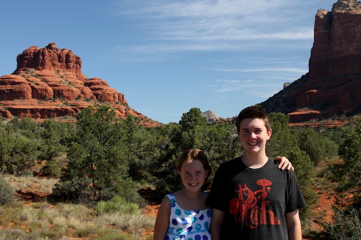 two people standing next to each other in front of red rocks and green trees on a sunny day