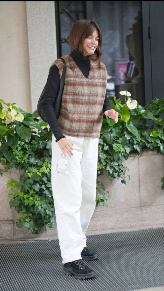 a woman standing in front of a building wearing white pants and a brown sweater with black sleeves