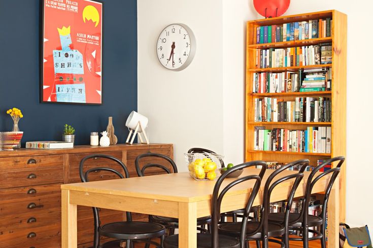 a dining room table and chairs in front of a bookshelf with a clock on the wall