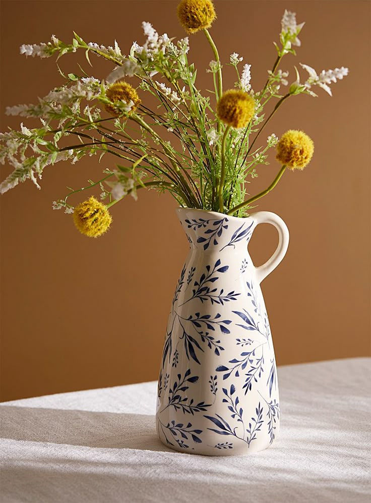 a blue and white vase with yellow flowers in it on a tablecloth next to a brown wall