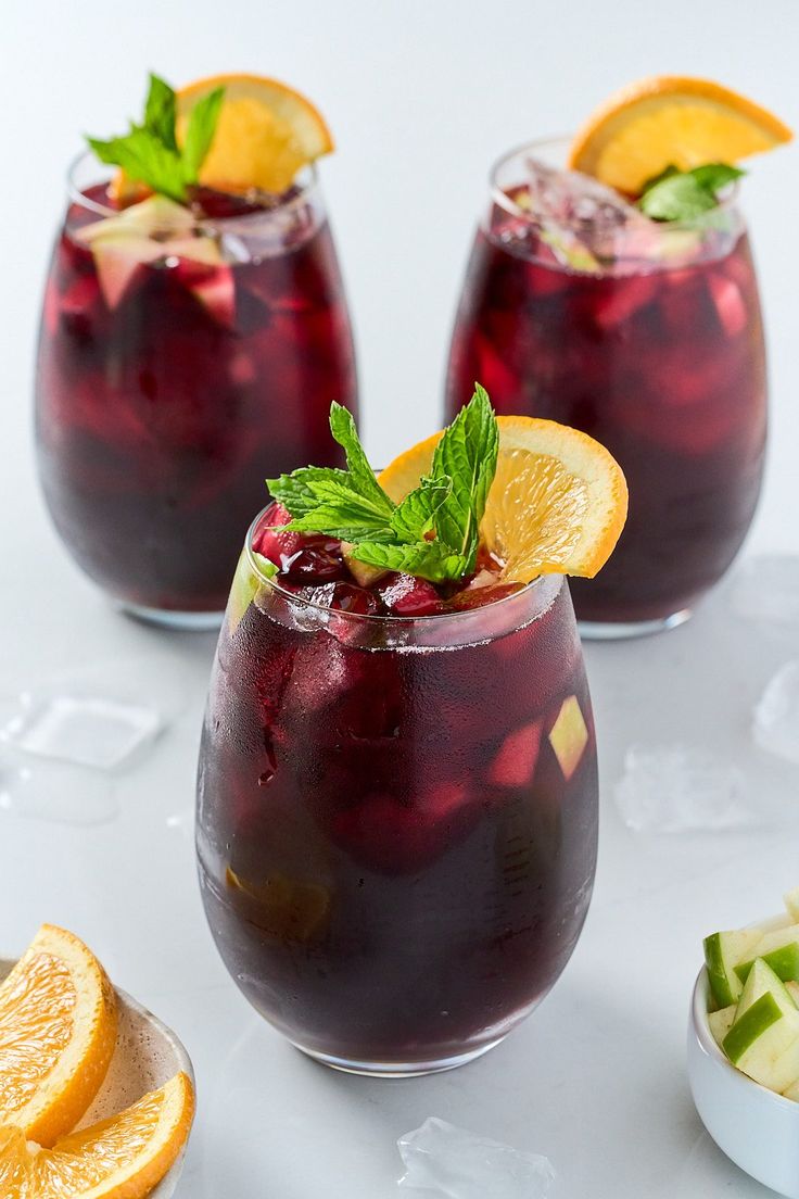 three glasses filled with red liquid and garnished with orange slices, mint and lemon wedges