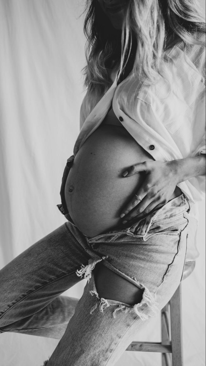 a pregnant woman sitting on a stool with her belly tucked under her shirt and ripped jeans