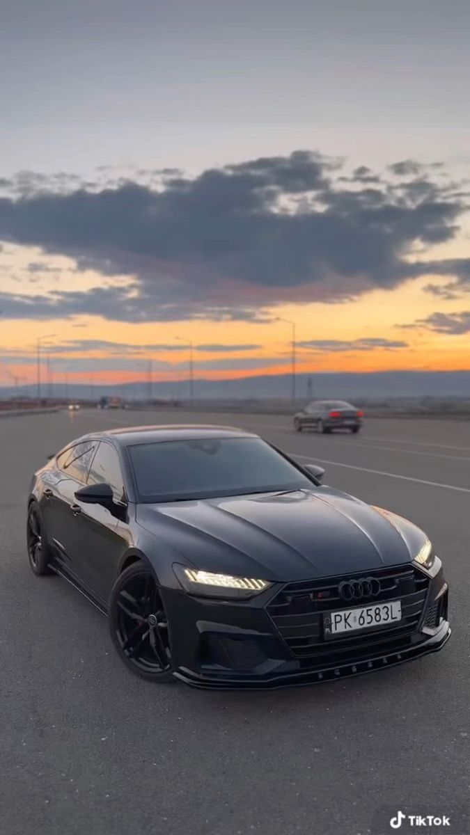 a black car is parked in an empty parking lot at dusk with the sun setting behind it