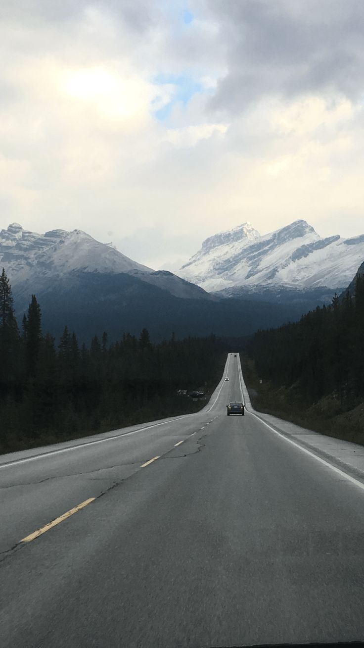 an empty highway with mountains in the distance and trees on both sides, under a cloudy sky