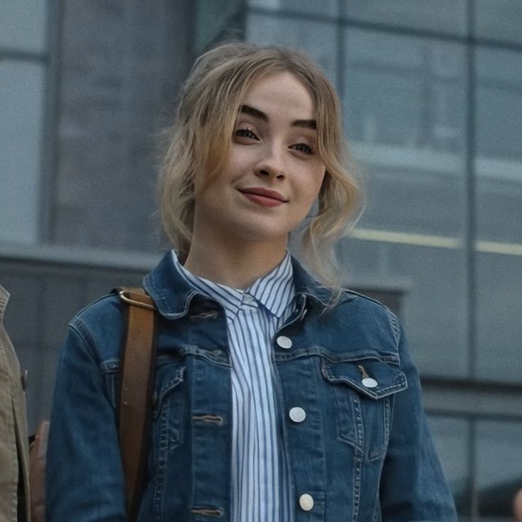 a woman with blonde hair wearing a denim jacket and carrying a brown bag in front of a building