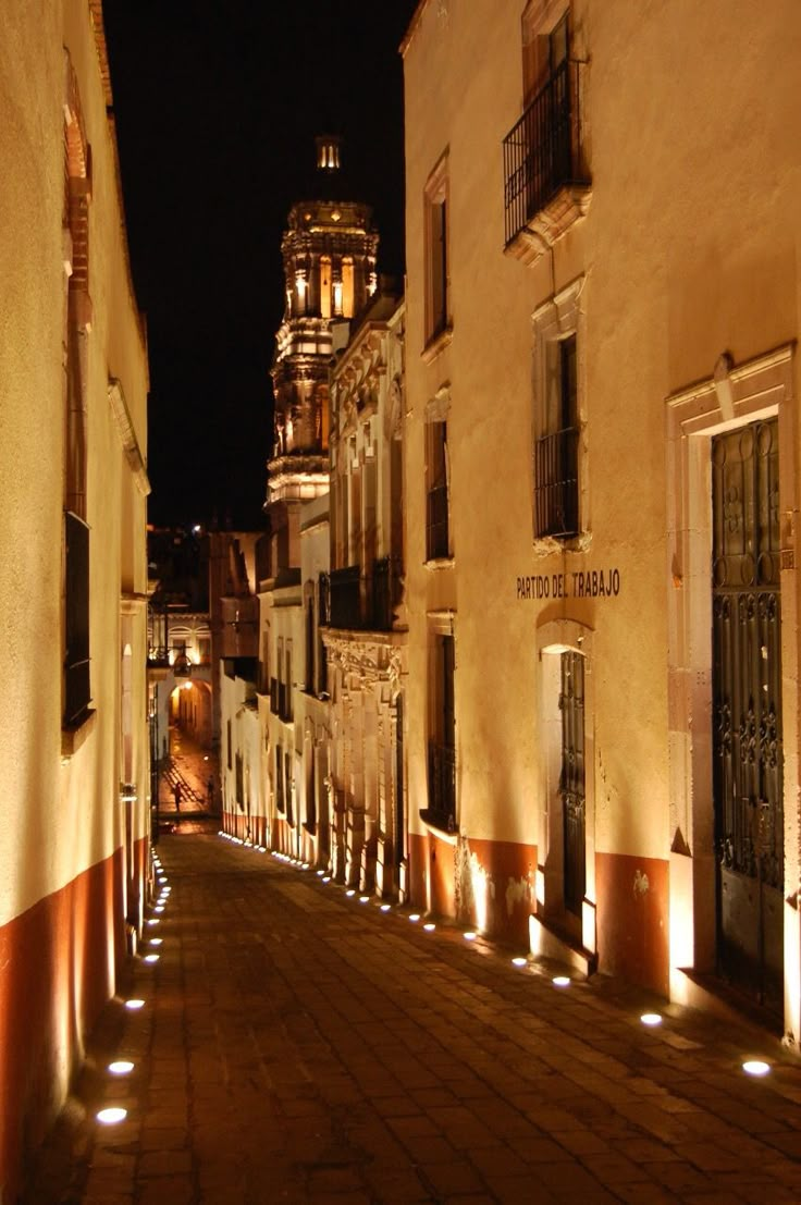 an empty street at night with lights on the buildings and door to another building in the distance