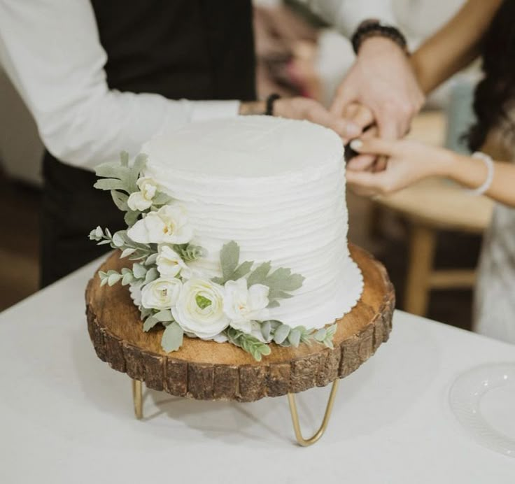 a man and woman cutting into a white cake on top of a wooden slice with greenery