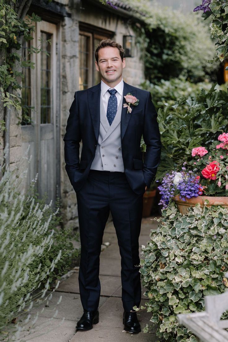 a man wearing a suit and tie standing in front of some flowers with his hands in his pockets