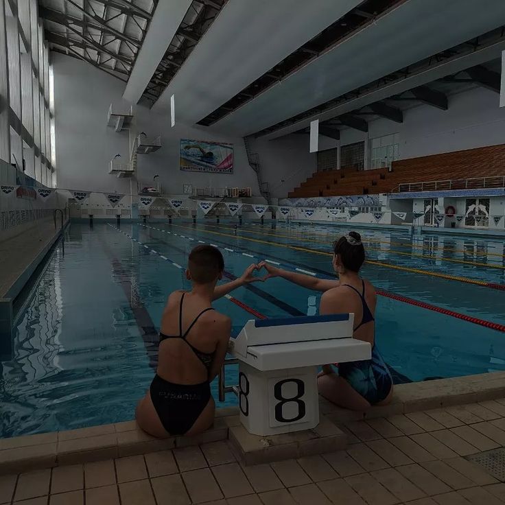 two women in bathing suits sitting on the edge of a swimming pool next to an electronic clock