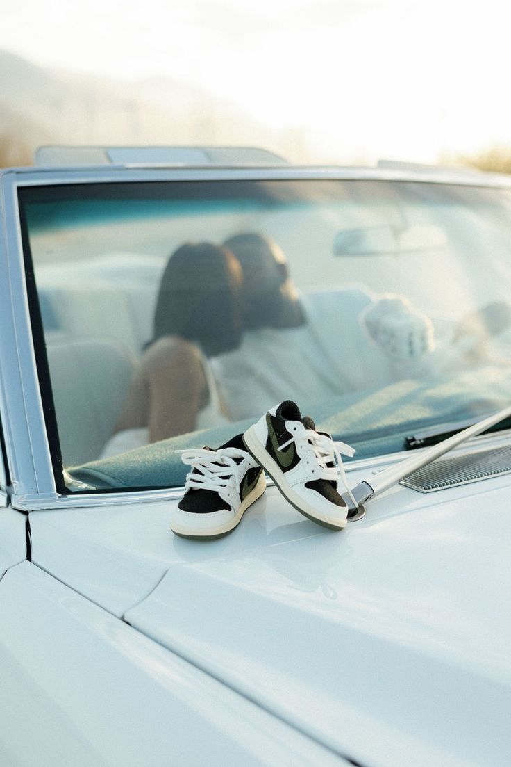 a pair of sneakers sitting on the hood of a white car with a woman in the background