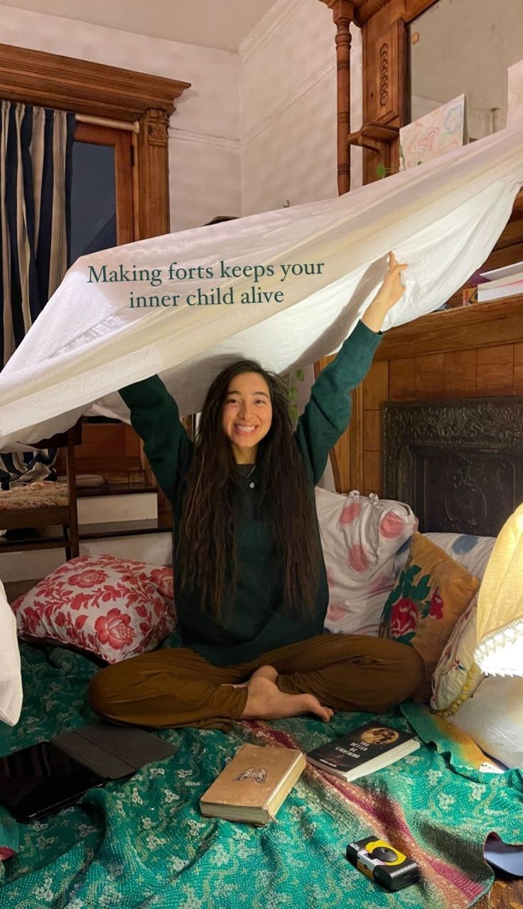 a woman sitting on top of a bed holding up a sheet over her head with the caption making forts keep your inner child alive