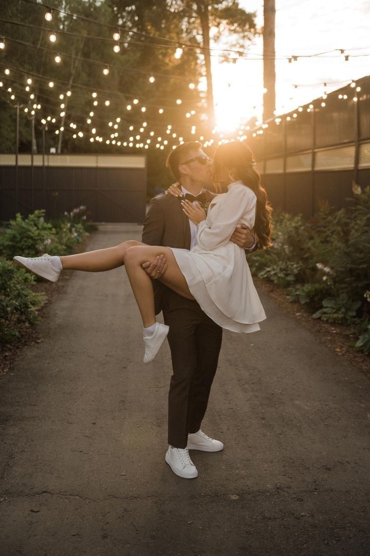 a man holding a woman in his arms as the sun sets behind them and string lights strung across the sky