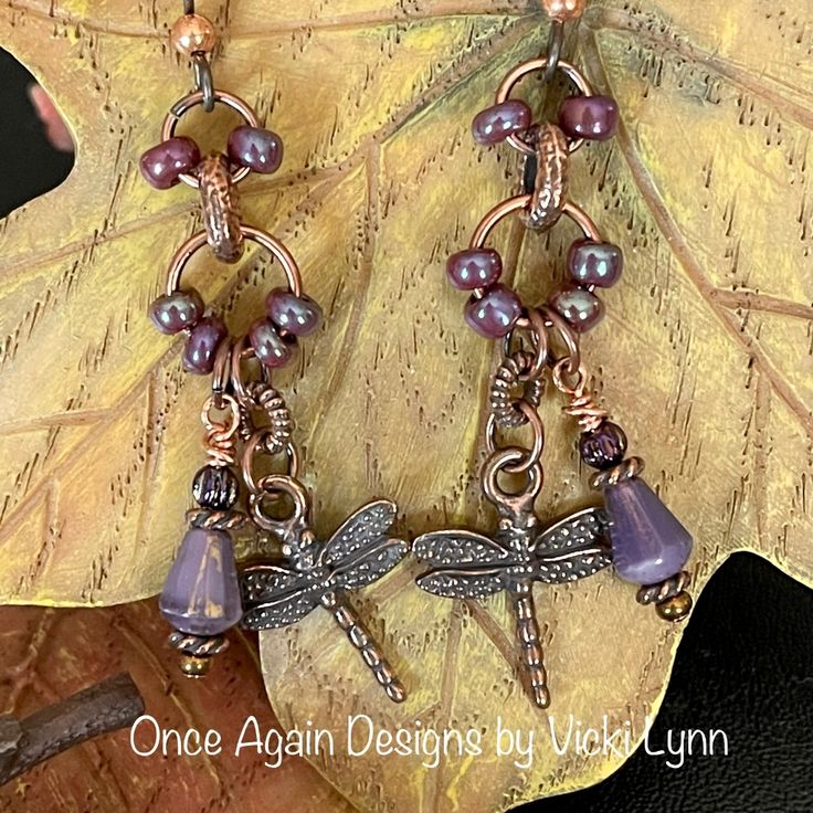 a pair of earrings with purple beads and dragonflys on them, sitting on top of a leaf