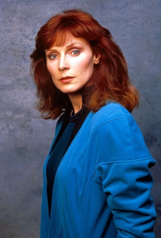 a woman with red hair and blue jacket posing for a photo in front of a gray background