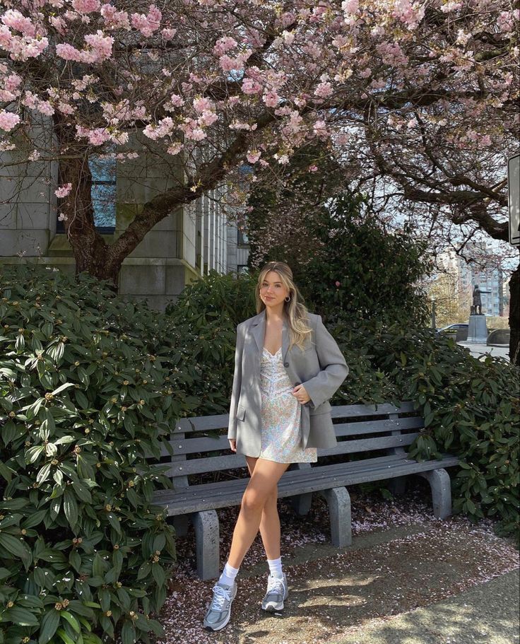 a woman is sitting on a bench in front of some bushes and trees with pink flowers