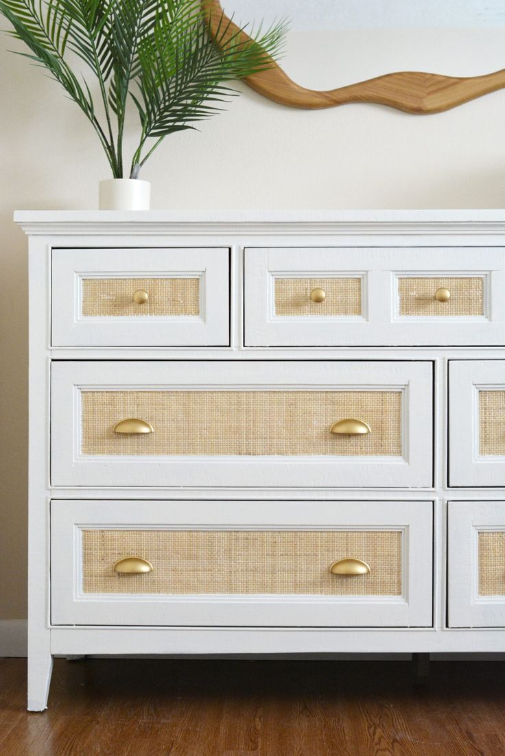 a white dresser with wicker drawers in front of a plant on top of it