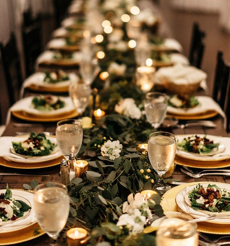 a long table is set with white plates and gold place settings, candles and greenery