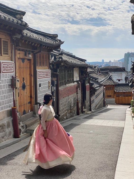 a woman in a pink and white dress is walking down the street with buildings behind her