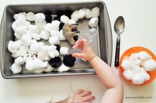 a toddler playing with cotton in a metal tray and spoons on the table