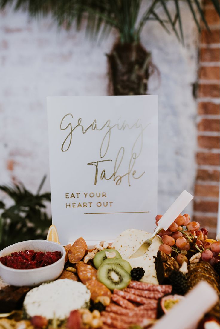 a table topped with lots of food next to a sign that reads grazing table eat your heart out