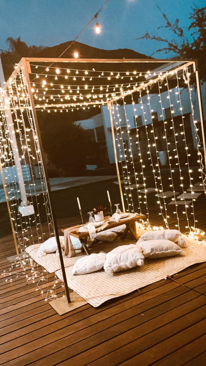 an outdoor dining area with string lights and pillows on the floor, surrounded by wooden decking