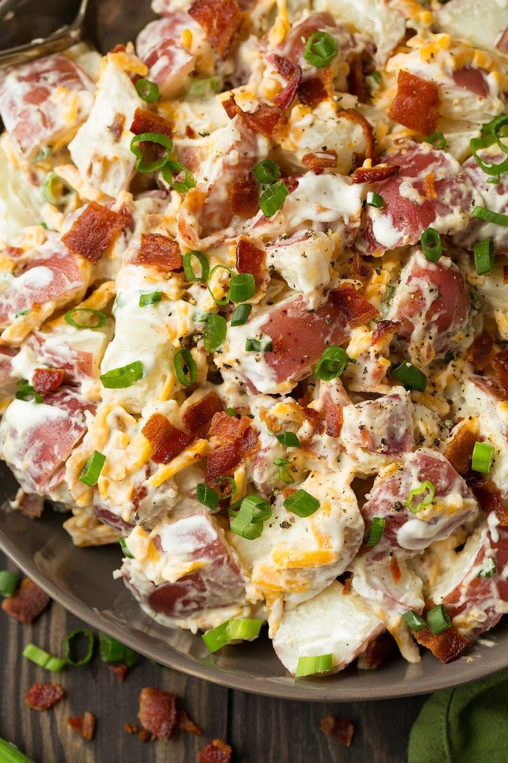 a bowl full of potato salad with bacon and green onions on the side next to a spoon