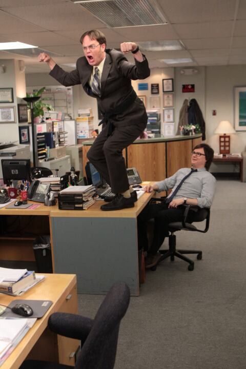 a man in a suit is jumping on a desk with two other men sitting around