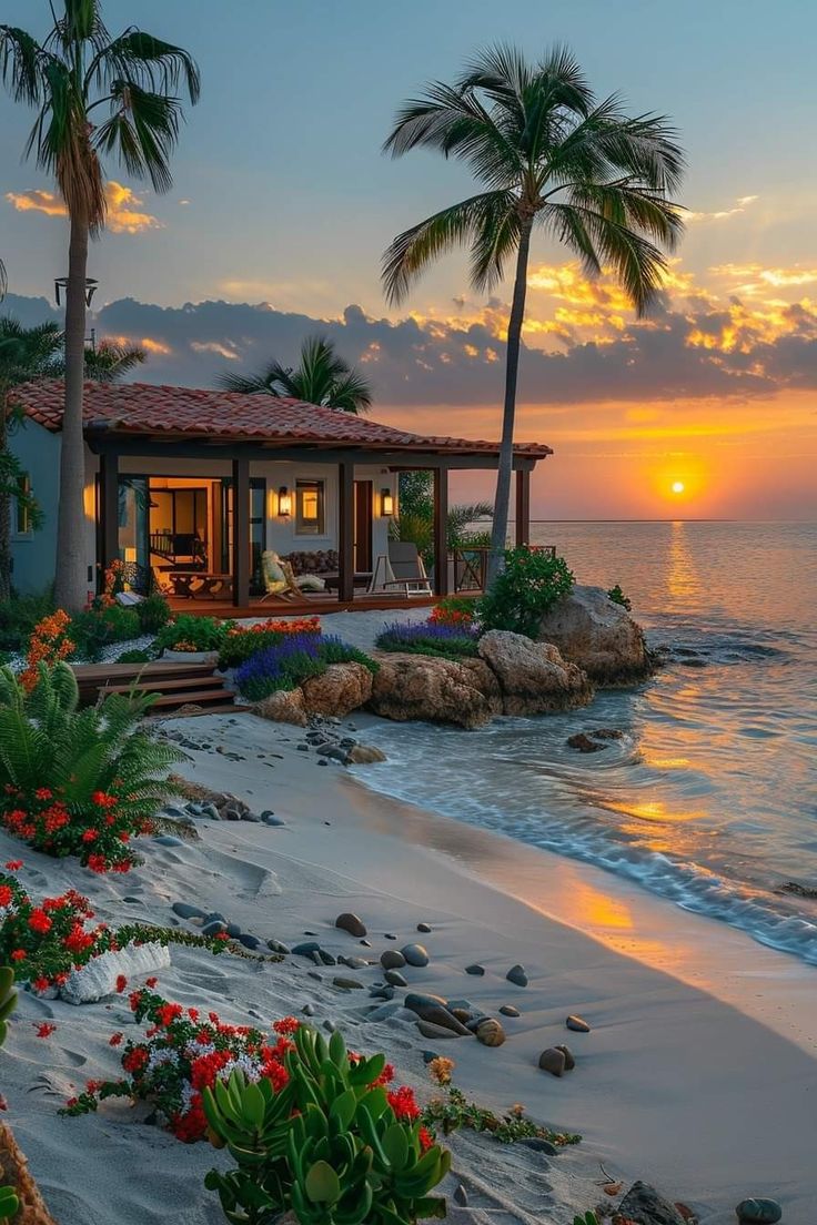 a house on the beach at sunset with palm trees and flowers in front of it