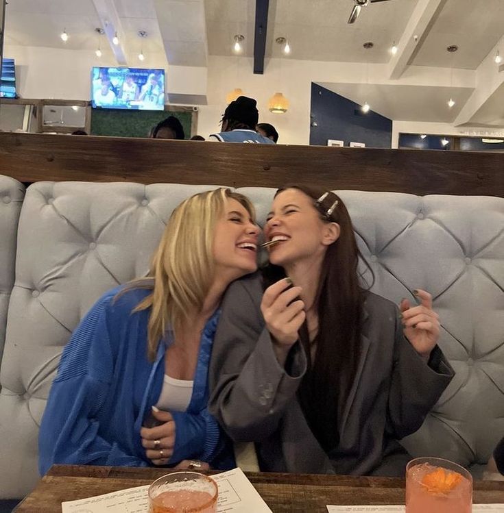 two women sitting at a table with drinks and papers in front of them, one is kissing the other's cheek