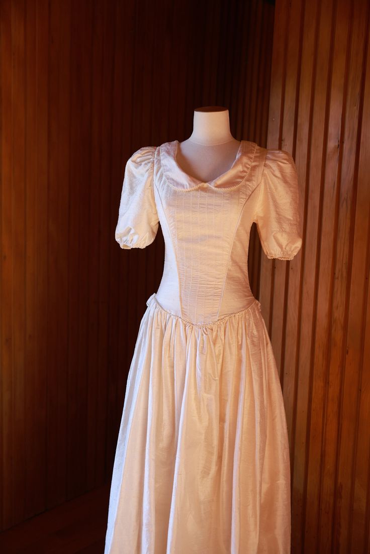 This silk dupion gown is an ode to 1980's lavish embellishment.  Unbranded but professionally made gown features a sweet Peter Pan collar with pearl edging, gorgeous puffy sleeves and a pin-tucked bodice shaped in a low vee. A gathered skirt with an extra train bustle at the back, edged in pearl trim and finished off with fabric rosettes. The condition of this dress is very good and would suit a petite bride. To fit: Bust 81cm Waist 57cm Hip 83cm Dress measures on mannequin: Shoulder to natural Fitted Puff Sleeve Dress For Debutante Ball, Silk Puff Sleeve Wedding Dresses, Vintage Satin Victorian Ball Gown Dress, Vintage Satin Victorian Ball Gown, Vintage Victorian Satin Ball Gown, Formal Gown With Fitted Bodice And Puff Sleeves, Elegant Victorian Satin Dress For Costume, Elegant Vintage Puff Sleeve Wedding Dress, Fitted Gown With Puff Sleeves For Wedding