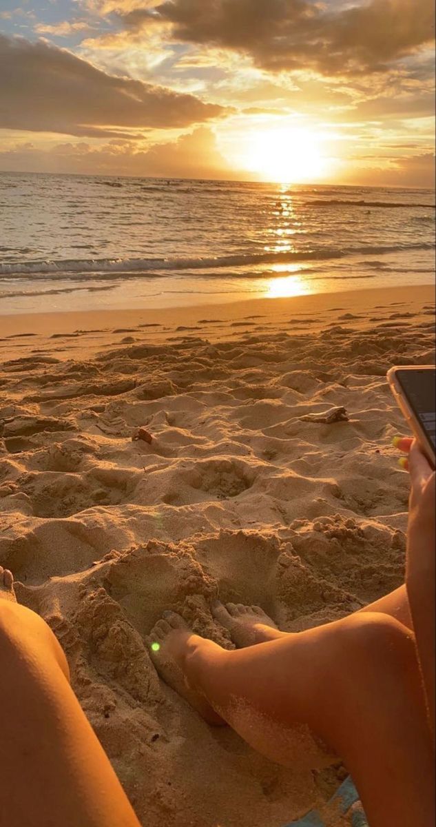 a person sitting in the sand with a cell phone on their lap looking out at the ocean