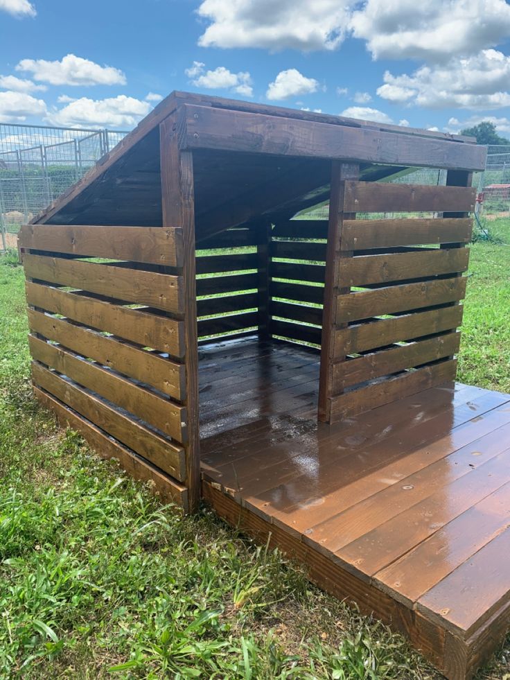 a dog house made out of pallets in the middle of a grassy field with blue sky and clouds